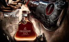 kompozice, která zachycuje dvě lahve prémiového koňaku Hennessy X.O. na dřevěném povrchu. V popředí je menší, elegantní lahev s jantarově zbarveným obsahem, vedle ní leží kovový uzávěr. V pozadí je větší, tmavší lahev s výrazným reliéfem a nápisem Hennessy. Na levé straně se nachází skleněná miska plná svinutých doutníků, což navozuje atmosféru luxusu a vytříbeného vkusu.