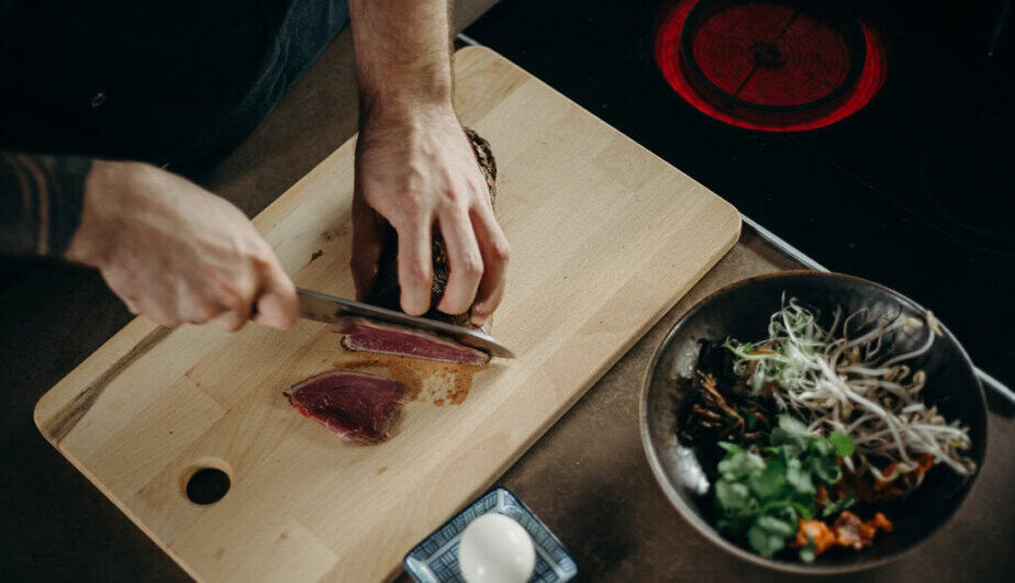 Ruce člověka krájejí maso ostrým kuchyňským nožem na dřevěném prkénku - ilustrační obrázek k článku, jak vybrat kuchyňský nůž na Trendymagazin.cz